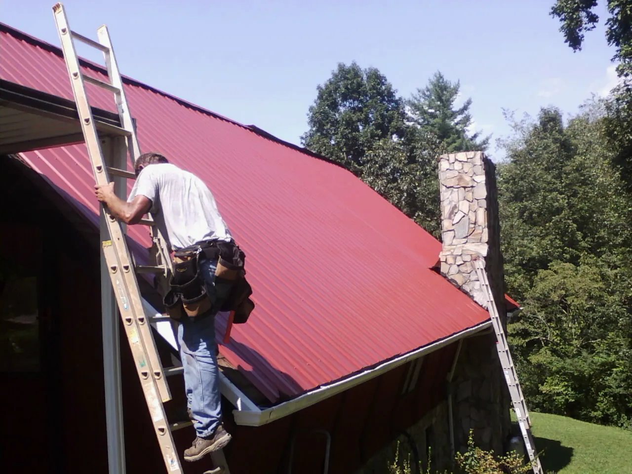 Roofer on a ladder beside a red metal roof and stone chimney — Soffit & Fascia Repair by Roofing Brothers in Myerstown